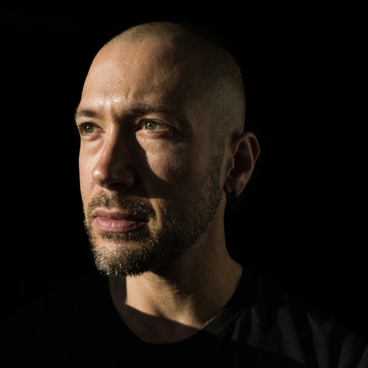 Color photo of a tanned, middle-aged Jew with a short graying beard and buzz cut hair, and gray-blue eyes. He’s dramatically lit from the side with a black background, and he gazes resolutely into the distance like he’s planning to climb a mountain or something.